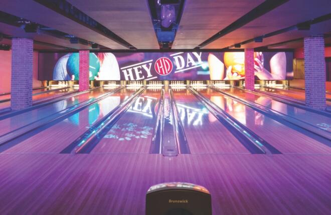 Brunswick bowling lanes at HeyDay Lower Bricktown in Oklahoma City, lit in purple and pink, with large LED screens above the pins displaying the HeyDay logo.