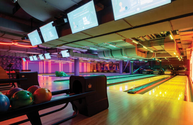 Modern bowling alley with neon lighting, bowling lanes, & colorful balls on rack