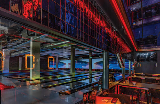 Stylish bowling lanes viewed through glass with dramatic red lighting and lane-side dining in an upscale bowling venue.