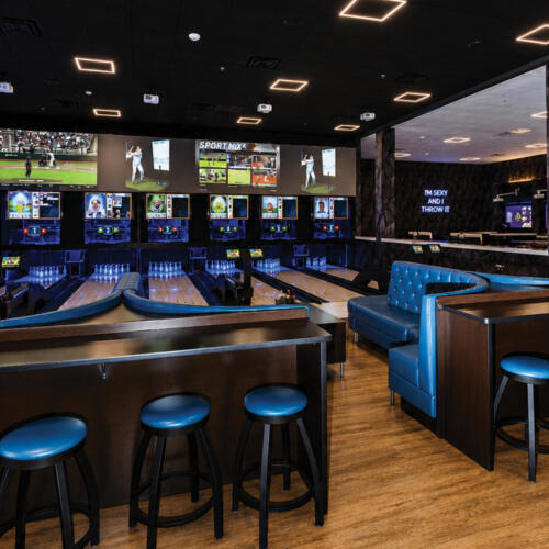 An angled view of the Crush It! bowling lounge showing blue leather bar stools and booths along wooden high-top tables, illuminated lanes with sports screens overhead, the "Let The Good Times Roll" neon sign on the left, and an "I'm Sexy and I Throw It" neon sign near the arcade in the background.