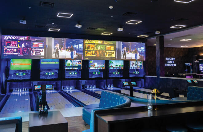 A wide view of the Crush It! bowling lounge with brightly lit lanes numbered 1 through 6, a large wall of sports and gaming screens, blue tufted leather booths, and neon "Let The Good Times Roll" and "I'm Sexy and I Throw It" signs on opposite walls.