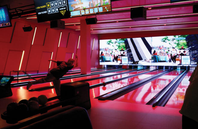 A bowler mid-release on a red-lit lane, with overhead score monitors displaying a "HORSE" game and Angry Birds, and a large LED wall at the lane's end showing an outdoor market scene.