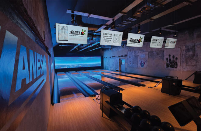 Perspective view down illuminated bowling lanes with overhead screens and contemporary scoring systems.