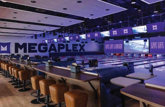 A modern bowling alley at Megaplex with rows of lanes under a large "MEGAPLEX" wall graphic, overhead screens reading "LIVE LIFE LARGER," and tan leather bar stools lining high-top tables in the foreground.