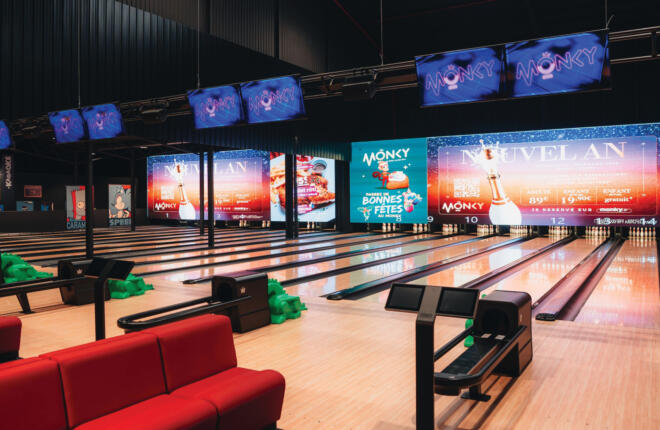 Wide view of contemporary bowling lanes with overhead scoring displays, branded digital backwalls, and red lounge seating.