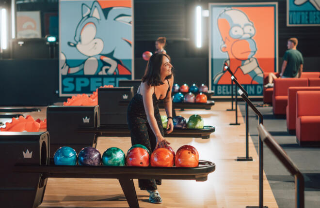Guest selecting a bowling ball beside contemporary lanes with vibrant seating and graphic wall art.