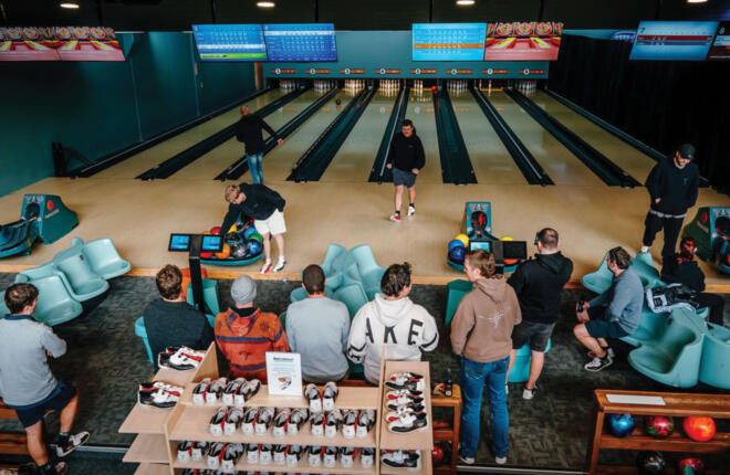 Busy bowling center with guests bowling, social seating, ball returns, and digital scoring screens overhead.