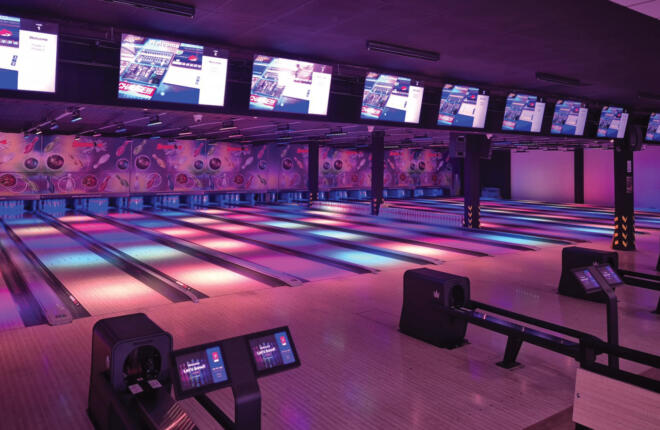 Neon-lit bowling lanes with overhead scoring screens and colorful LED lane lighting at Strike Yard Bowling.
