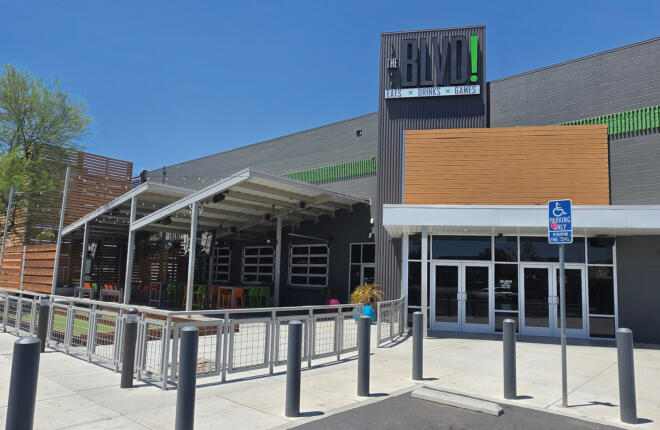 Daytime exterior of The BLVD! showing the gray building with wood-slat accents, a covered patio with colorful seating, and the illuminated "THE BLVD! Eats × Drinks × Games" tower sign against a clear blue sky.