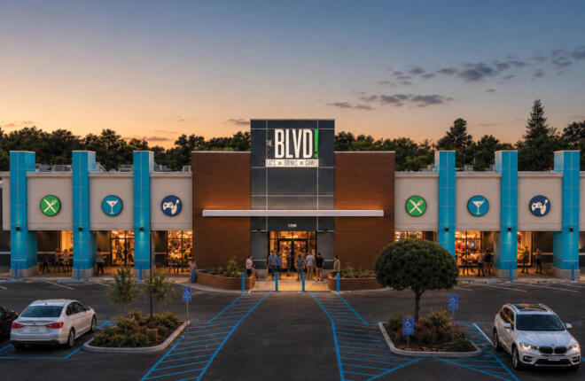 Exterior of The BLVD! at dusk, showing the brick and blue-tiled storefront with the illuminated "THE BLVD! Eats × Drinks × Games" sign, entrance with people gathered, and cars in the parking lot.