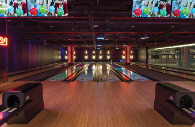 Bowling lanes with wooden floors, large screens, neon signs, industrial ceiling.