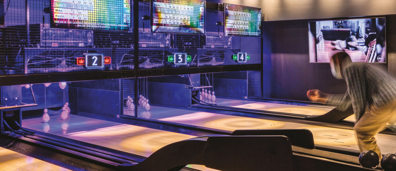 A player releases a ball down a duckpin lane with overhead digital scoreboards and hanging string pins visible under purple lighting.