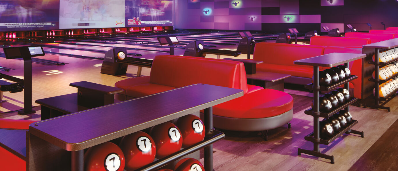 Bowling lanes with red lounge seating, black and white ball racks, and large overhead screens.