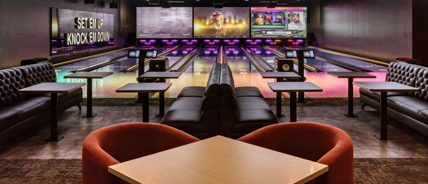 Dark leather lounge seating and tables facing lit bowling lanes with large display screens above.
