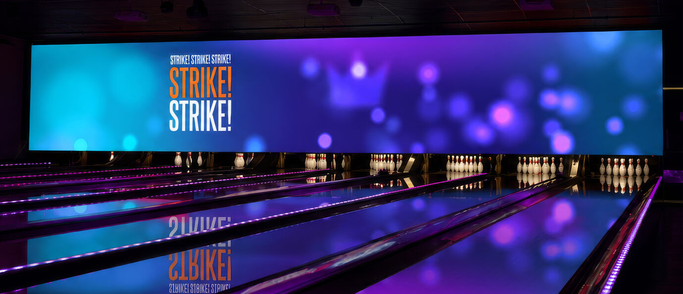 Brunswick LED Video Wall displaying bold "STRIKE! STRIKE! STRIKE!" typography in orange and white over a blue and purple bokeh light background, spanning the full width of the pin deck across multiple bowling lanes.