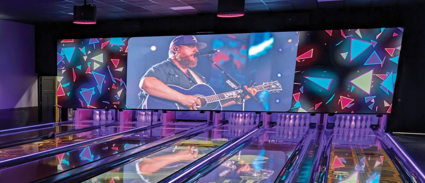 Brunswick LED Video Wall at Shawnee Lanes featuring a live concert performance of a country musician playing guitar, surrounded by colorful 80s-style geometric triangle graphics above a row of bowling lanes.