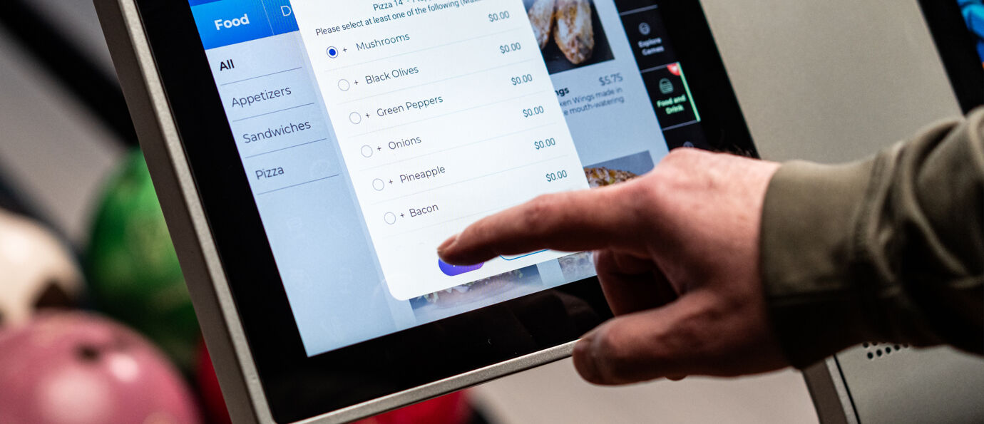 Guest selects pizza toppings from a Sync scoring tablet touchscreen, choosing from mushrooms, peppers, and more.
