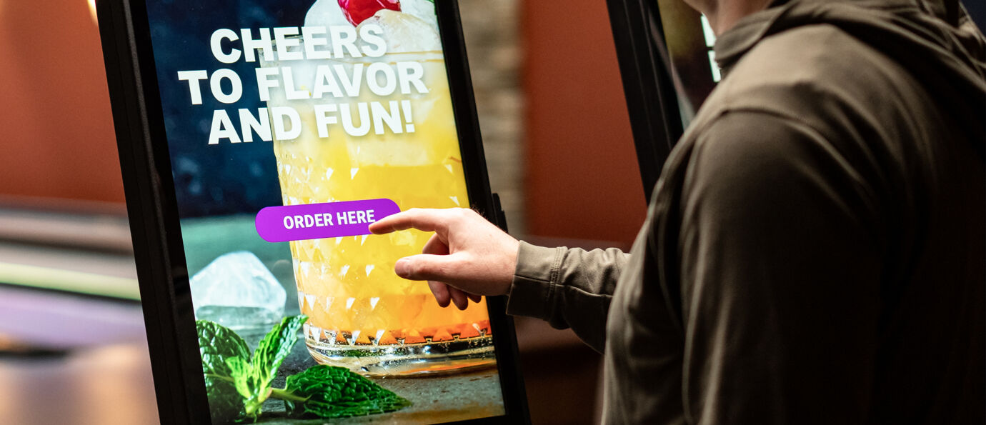 Guest taps "Order Here" on a Sync kiosk screen displaying a colorful cocktail with garnish.