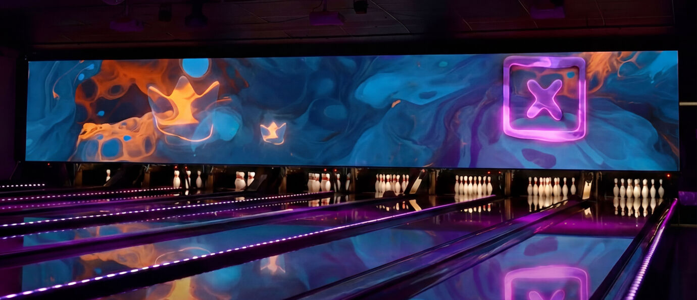 Brunswick LED Video Wall above bowling lanes displaying a vibrant blue and orange lava-flow motion graphic with a glowing neon crown and X logo, illuminating the pin deck and lane surfaces in purple tones.