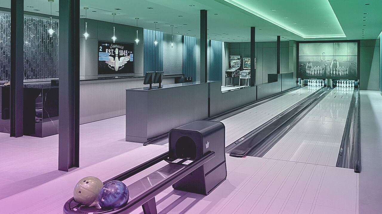 Luxury private bowling alley with multiple Brunswick lanes, ball return units, scoring consoles, pendant lighting, and green LED cove lighting. Brunswick bowling balls in the foreground; vintage bowling photography displayed on the back wall.