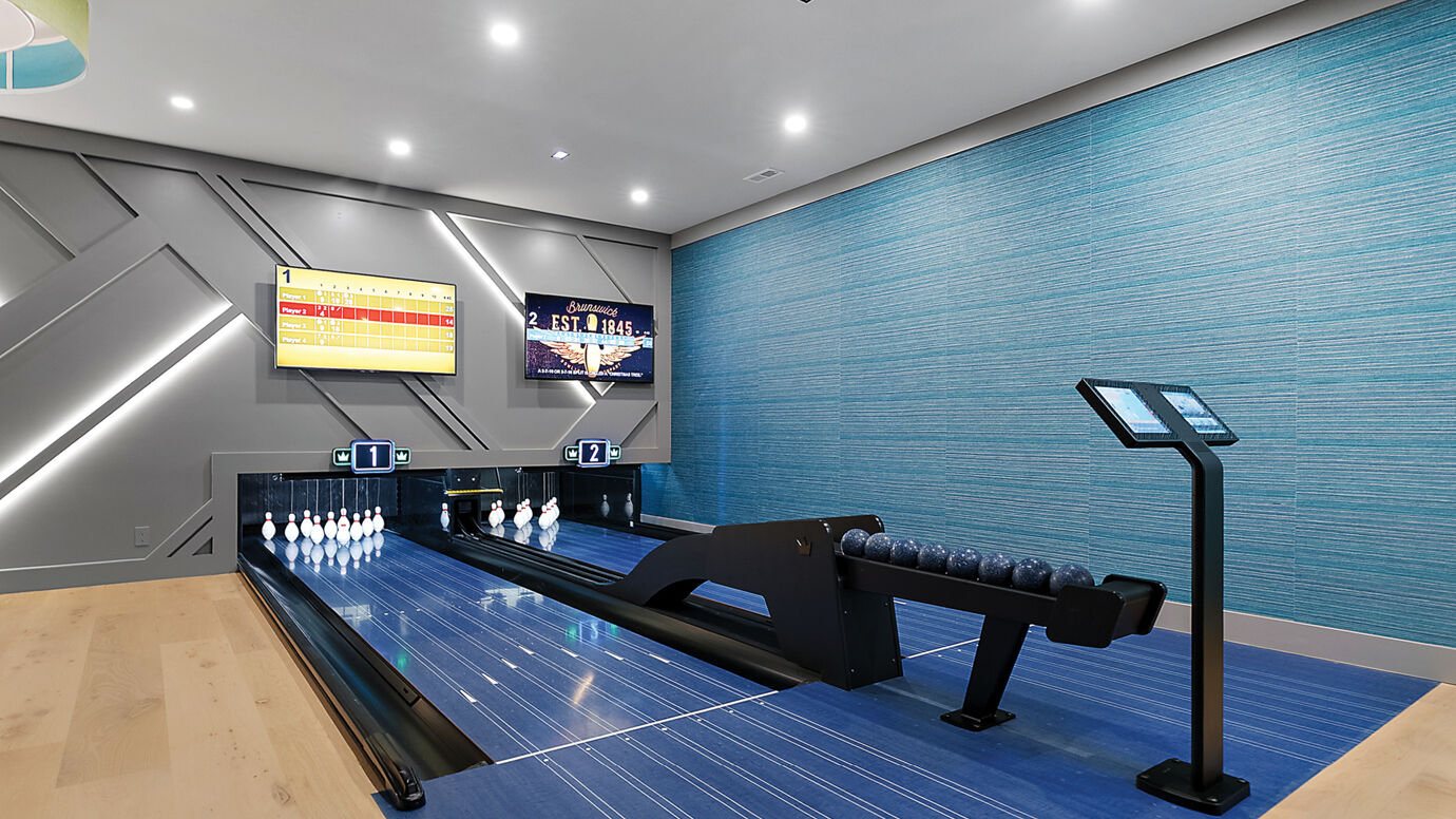 Close-up view of a two-lane residential bowling setup with Brunswick Sync scoring screens, illuminated pin decks, and sleek geometric wall lighting.